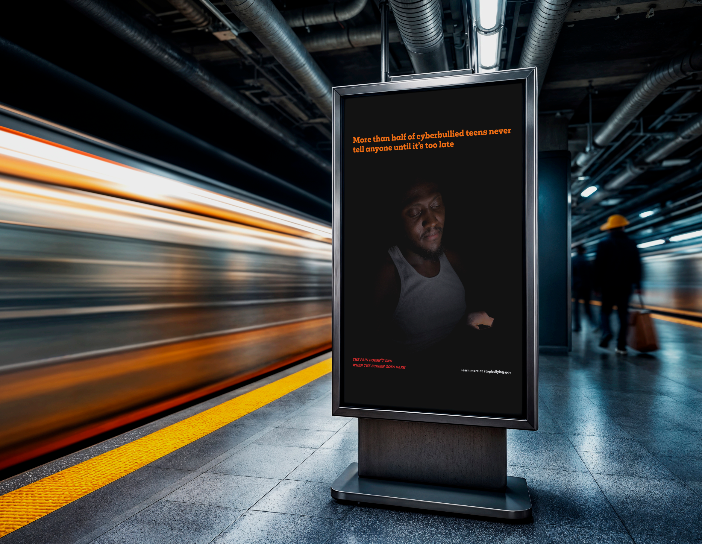 A digital billboard in a train station displaying a message about cyberbullying. The message states, 'More than half of cyberbullied teens never tell anyone until it’s too late,' alongside a photo of a sad young person looking down at a phone. The station has black ceilings with exposed pipes and a blurred train moving past.