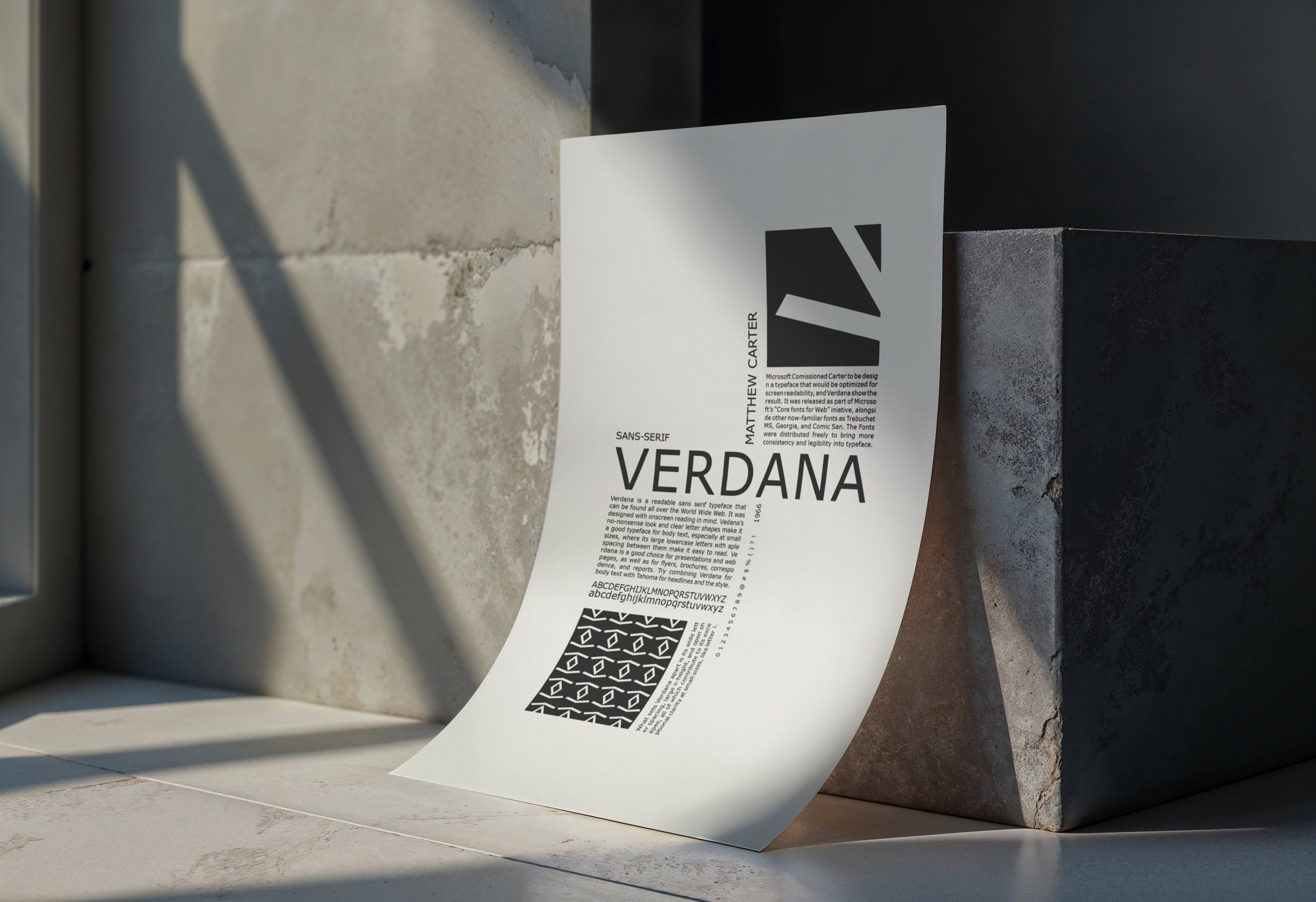 A piece of paper with text and graphics rests on a concrete surface near a brick wall. The paper displays the name 'Verdana' in large font, along with paragraphs of smaller text, and a black rectangle with white geometric patterns.