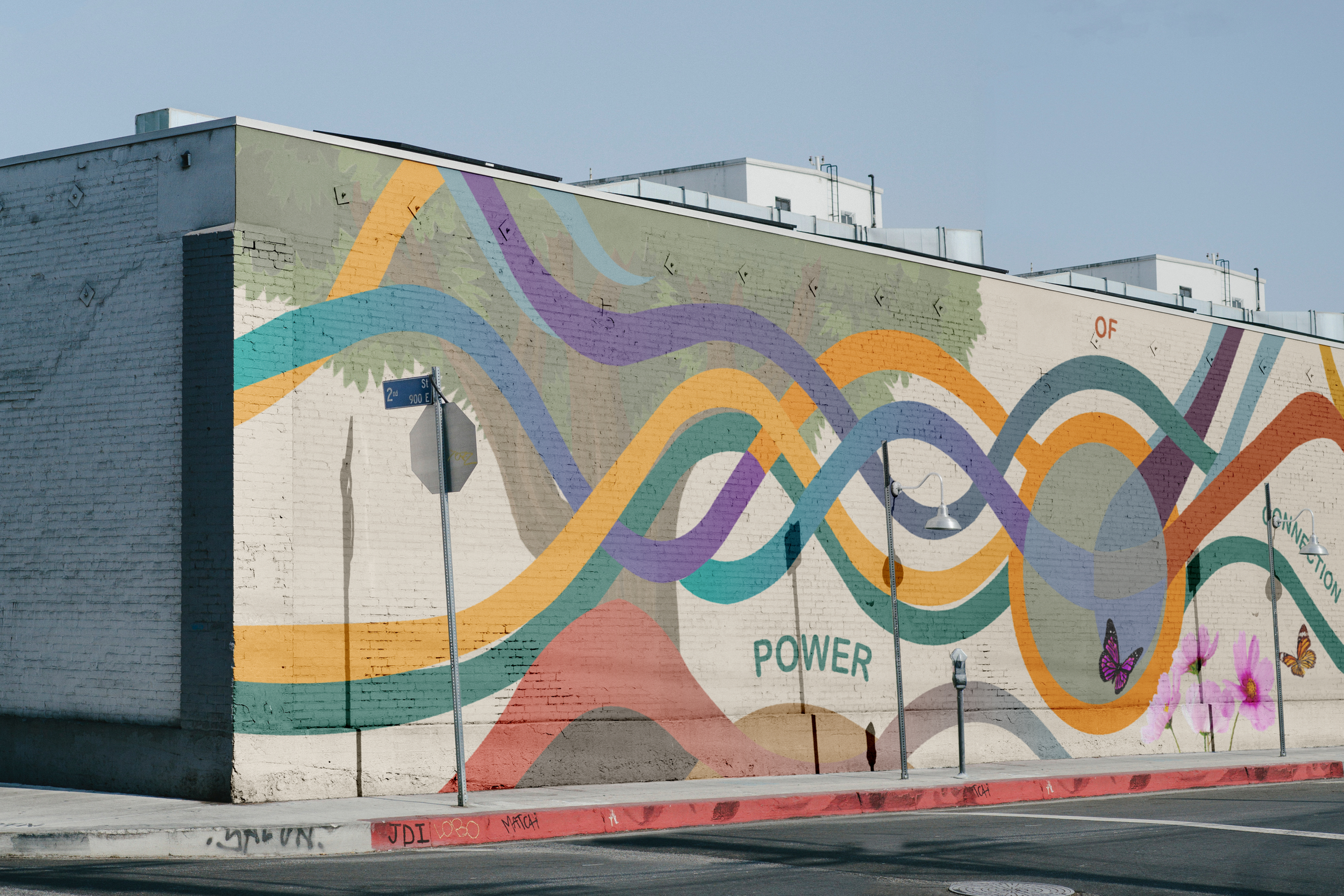 Colorful mural painted on a white brick building exterior, featuring swirling patterns and abstract designs in pastel shades of blue, purple, orange, green, and pink with some butterflies and flowers at the bottom.