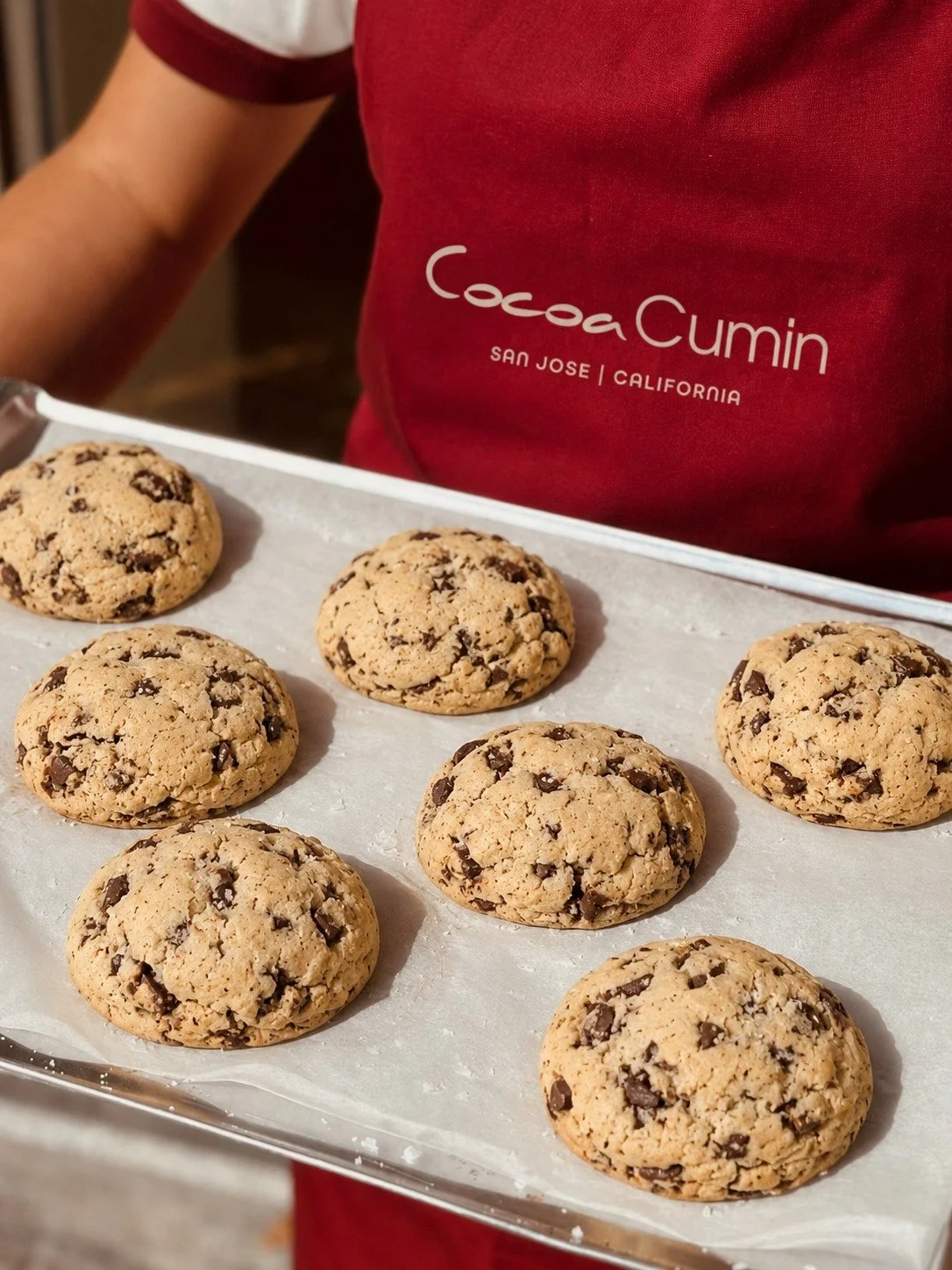 A tray of freshly baked chocolate chip cookies on parchment paper, with a person in a red shirt with 'Cocoa Cumin San Jose California' printed on it.