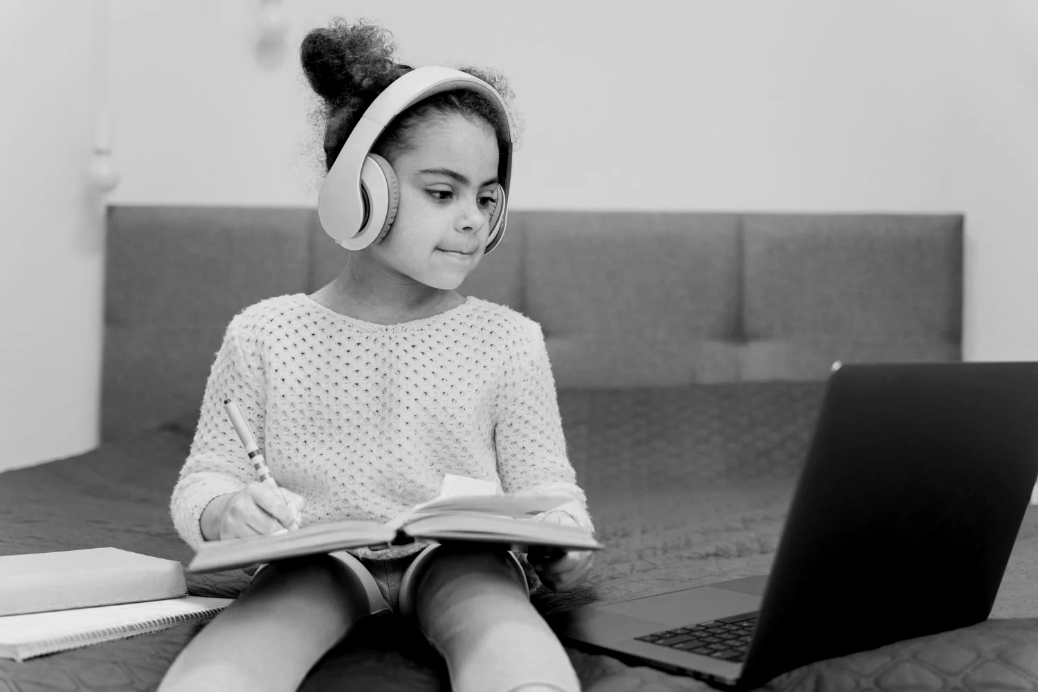 A young girl sitting on a bed, wearing headphones, reading a book, writing in a notebook, with a laptop nearby to depict online dyslexia tutoring