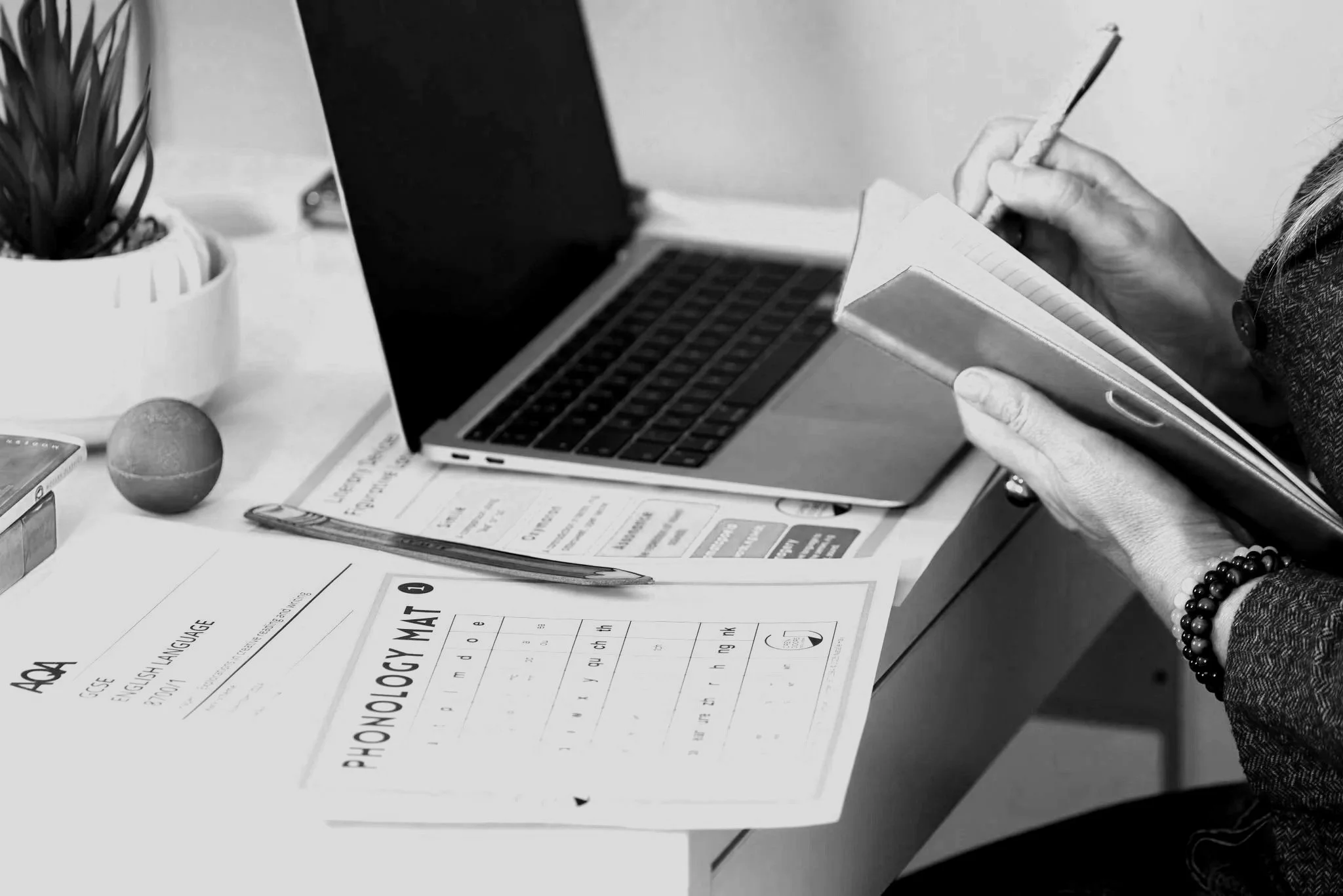A person's hand holding a pen and writing in a notebook on a workspace, with a laptop, papers, a potted plant, and a small ball on the desk.