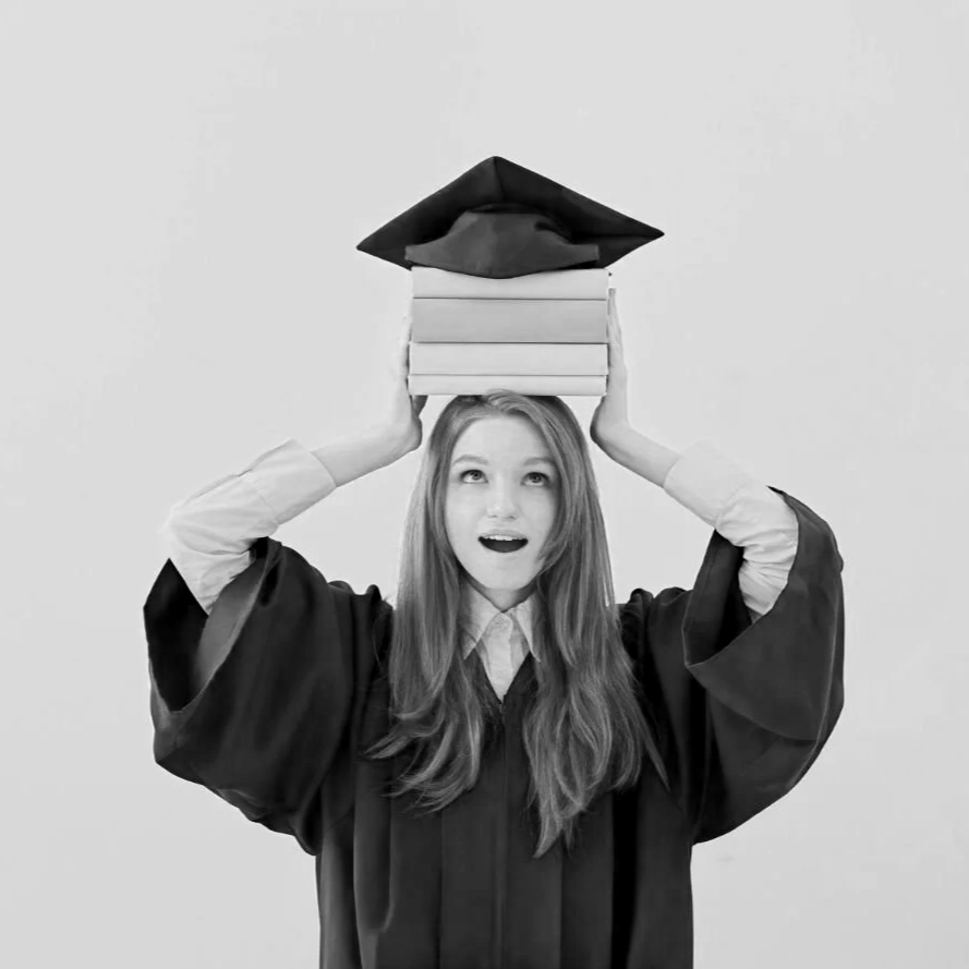 A teenager in graduation gown balancing a stack of books with a graduation cap on her head.