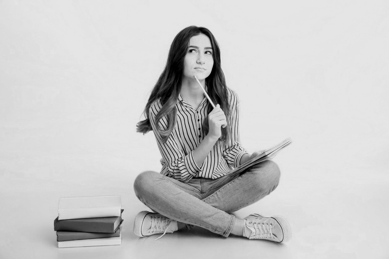 A young woman with long hair, sitting cross-legged on the floor, thinking with a pencil in her hand, holding an open notebook, with a stack of books beside her, against a plain background.