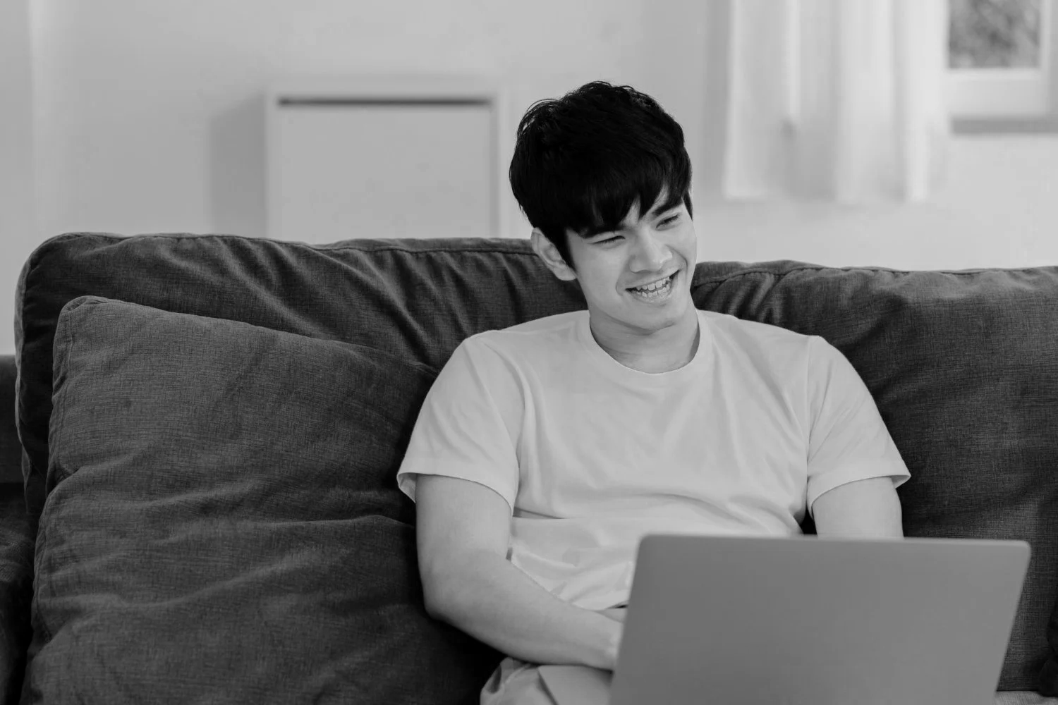A young man with dark hair, sitting on a couch with dark cushions, smiling and looking at a laptop to depict online dyslexia tutoring