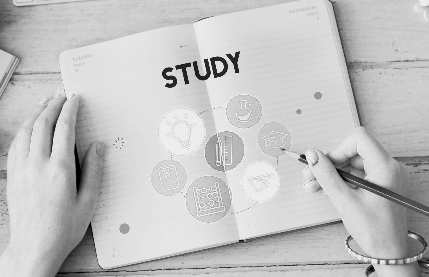 A person is writing in a notebook with the word 'STUDY' on the page, surrounded by icons related to learning and education, such as a lightbulb, flask, graduation cap, and paper airplane, on a wooden table.