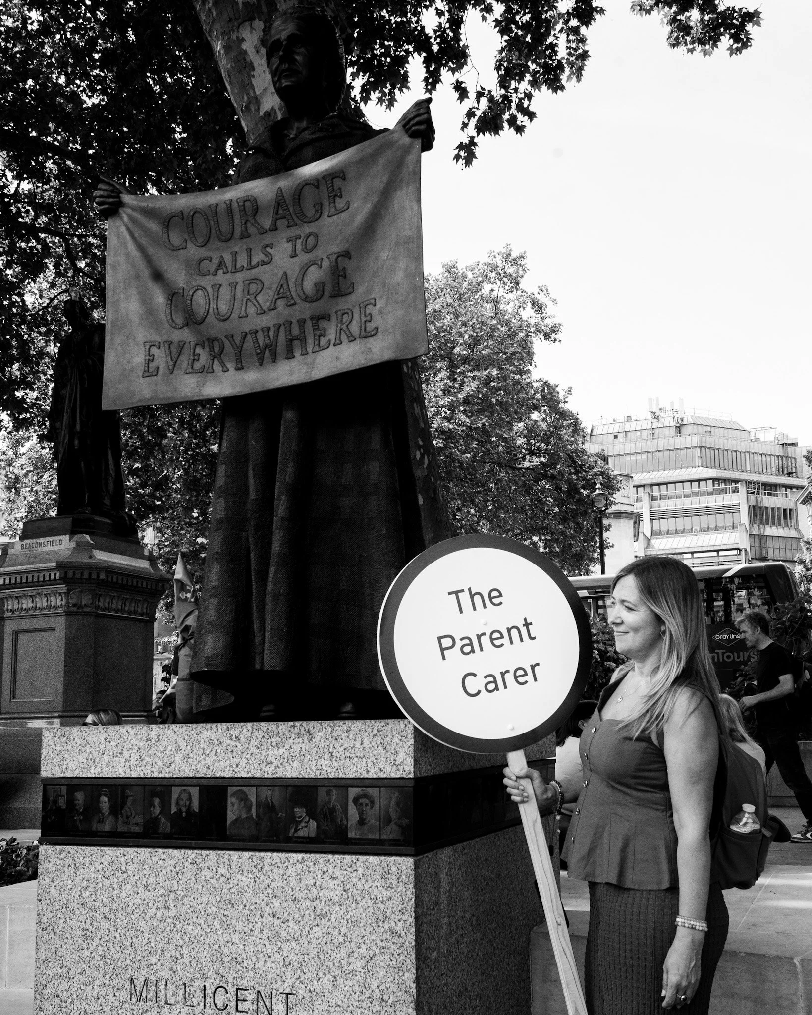 Natalie Hay holds a sign that says 'The Parent-Child Care' standing beside a large monument with a statue of a woman holding a banner that reads 'Courage Calls to Courage Everywhere'. The monument has portraits of various people at its base.
