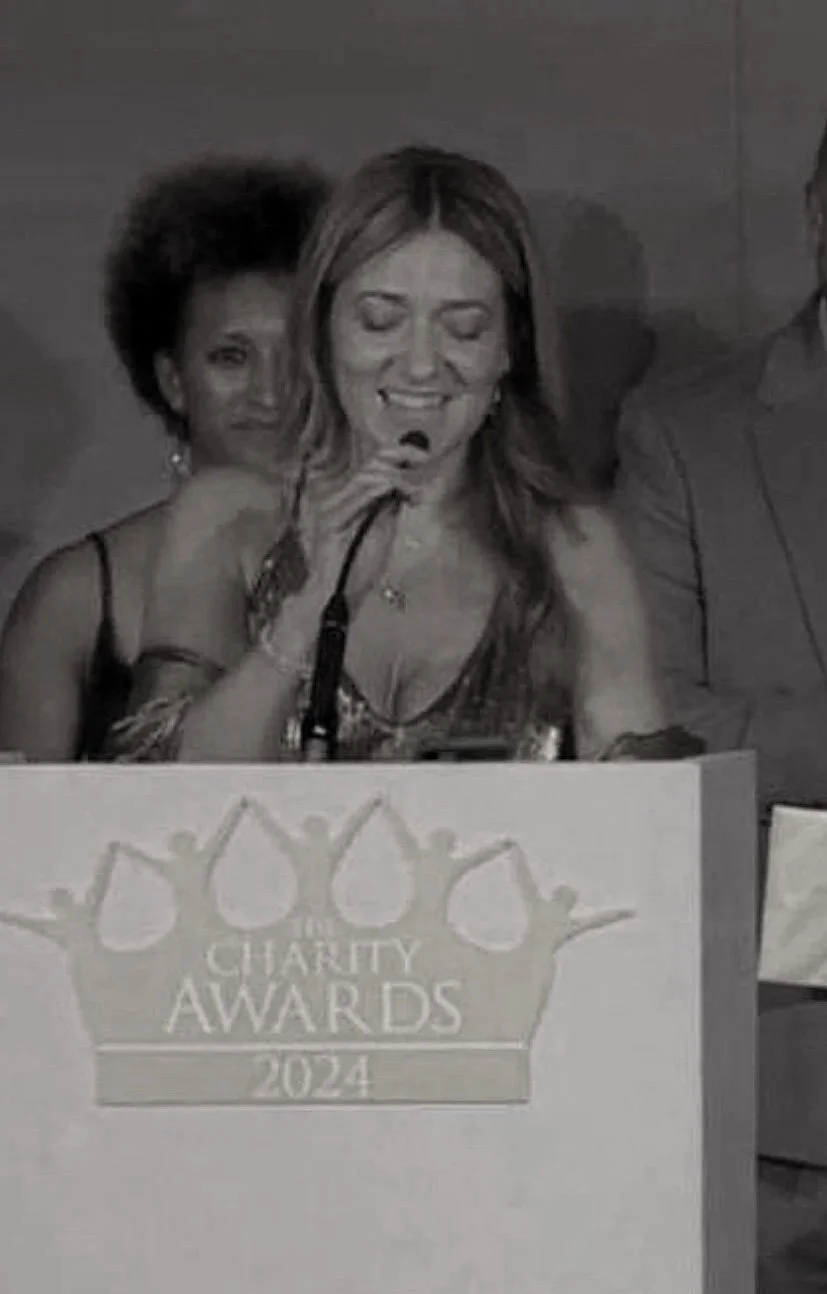 Natalie Hay speaking at a charity awards event, standing behind a podium with the words 'Charity Awards 2024' on it, while a woman in the background looks on.