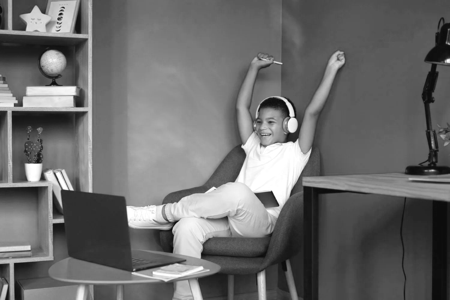 A boy sitting in a chair with headphones on, smiling and stretching his arms, with a laptop, tablet, and books nearby in a cozy room.