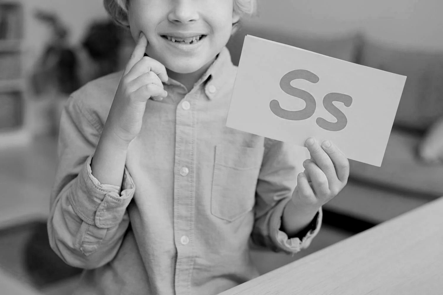 A young boy smiling and holding a card with the letter 'S' on it, while touching his face with his other hand, in a classroom setting.