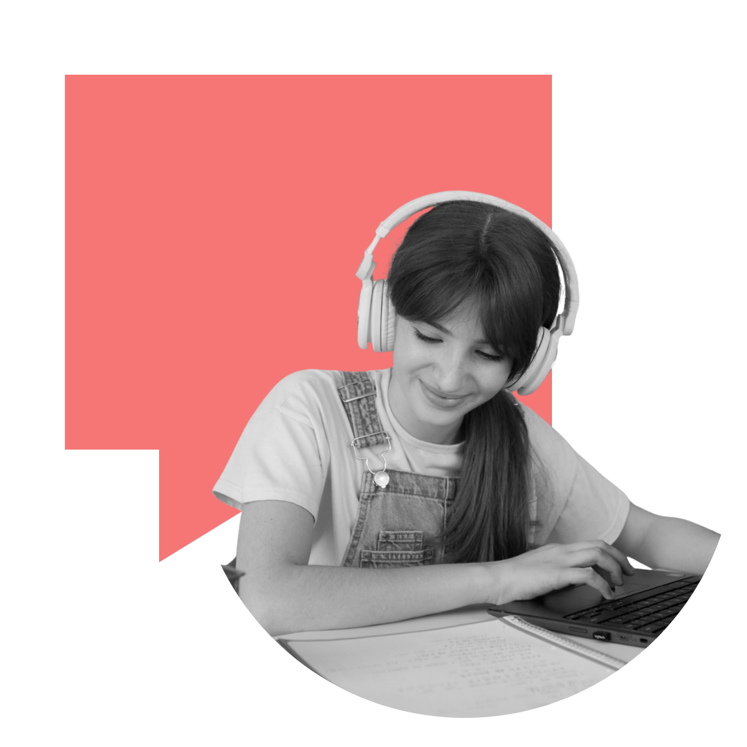 A young woman with long dark hair, wearing a T-shirt and denim overalls, smiling while using a laptop with headphones on, seated at a desk with an open notebook.