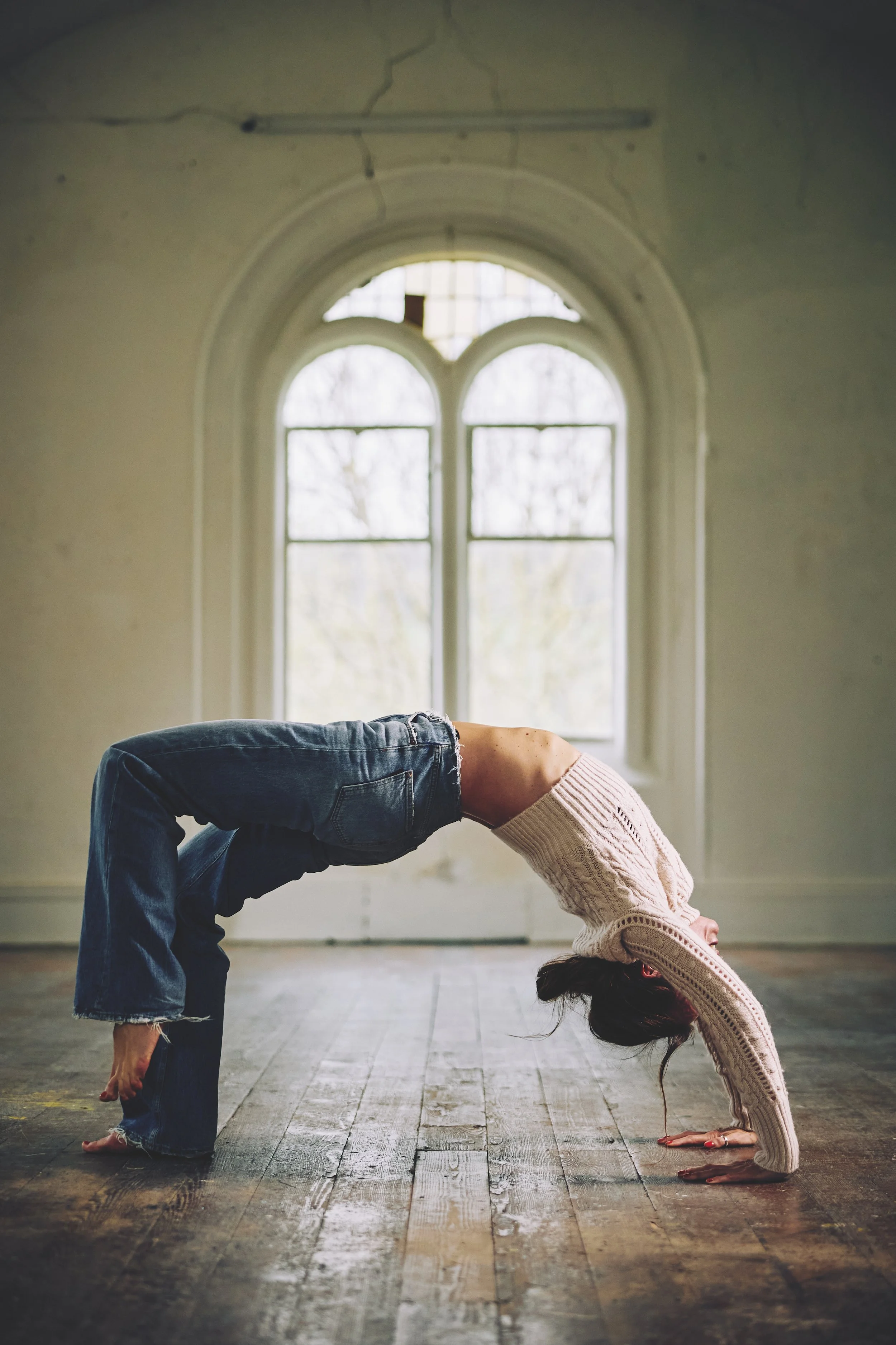 A woman practicing yoga in a spacious room with wooden floors and a large arched window, performing a backbend with her hands and feet on the ground and her body curved upward.