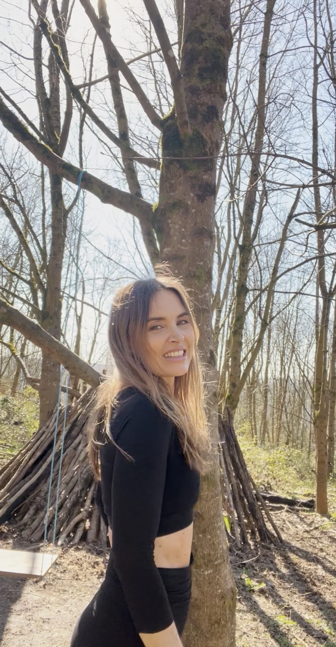 A woman smiling outdoors, standing in front of a tree with a teepee-like structure made of branches behind her. The scene is in a wooded area with leafless trees and sunlight.