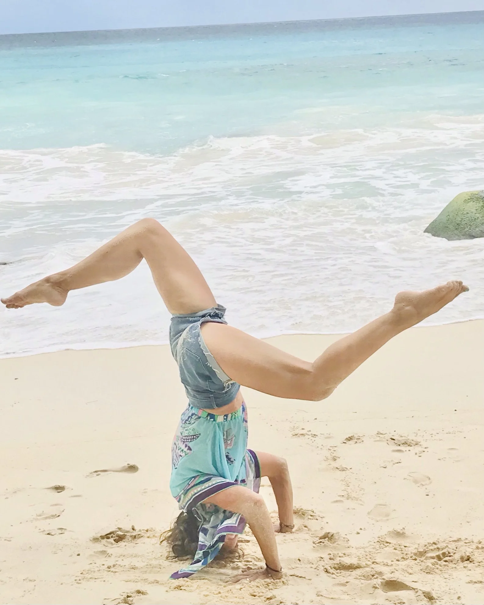 A person on the beach performing a handstand with legs bent and arms supporting their body, wearing shorts and sneakers, with the ocean and sky in the background.