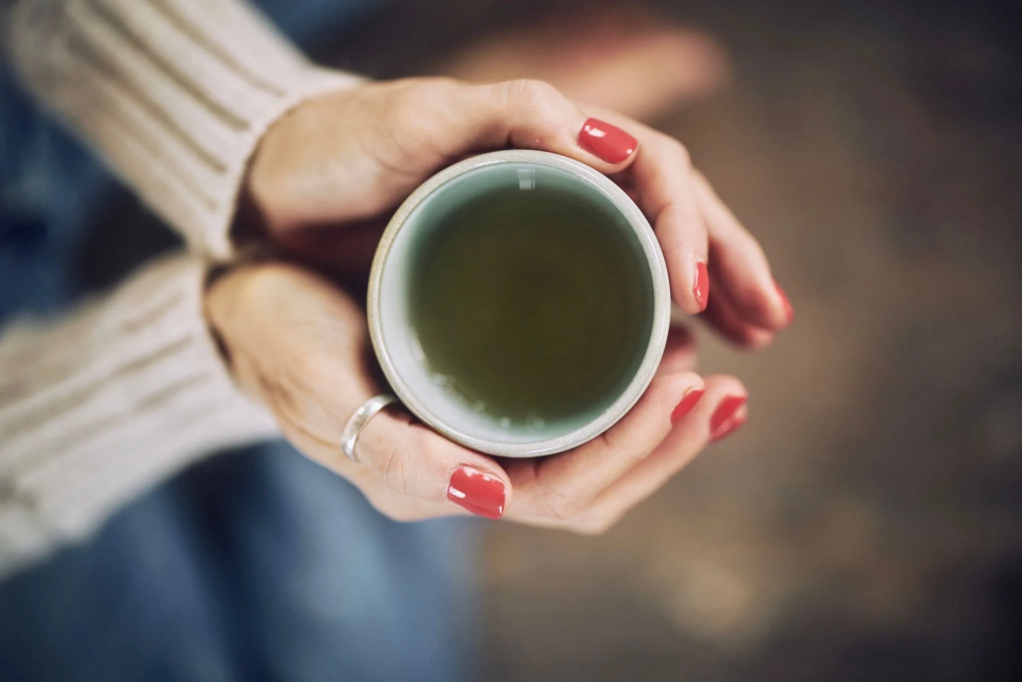 A person with red nail polish holding a white ceramic mug filled with a green beverage, wearing a cream-colored sweater.