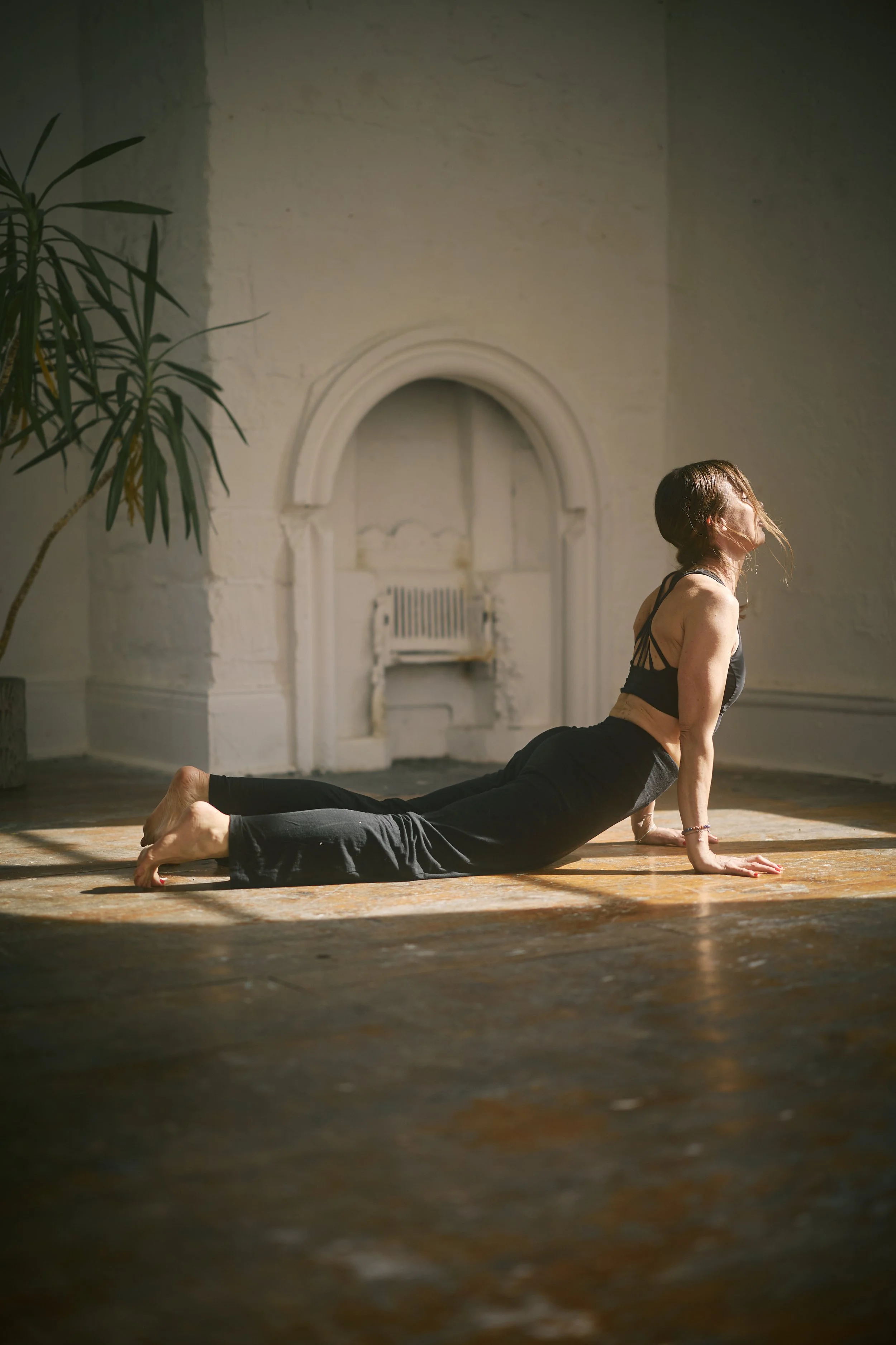 A woman practicing yoga in a sunlit room with a vintage wooden floor, an arched wall opening, a radiator, and a tall plant on the left side.