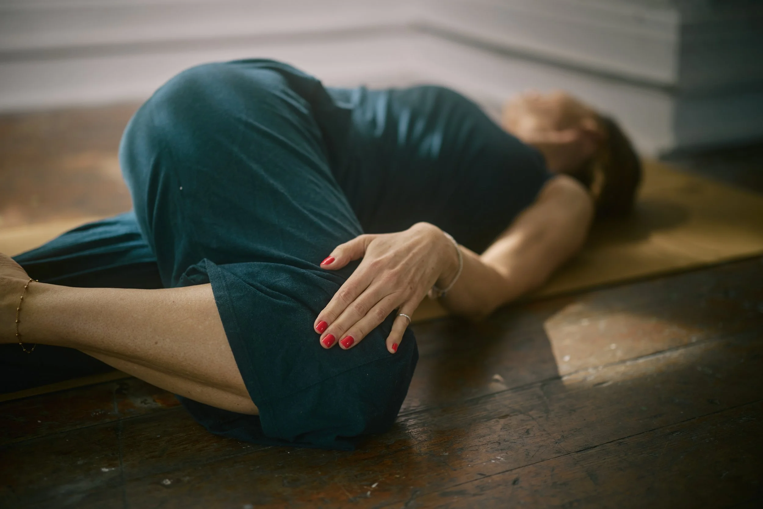 A person lying on the floor with crossed arms, wearing a black dress and dark green shorts, with red nail polish, on a wooden floor near a white wall and brick chimney.