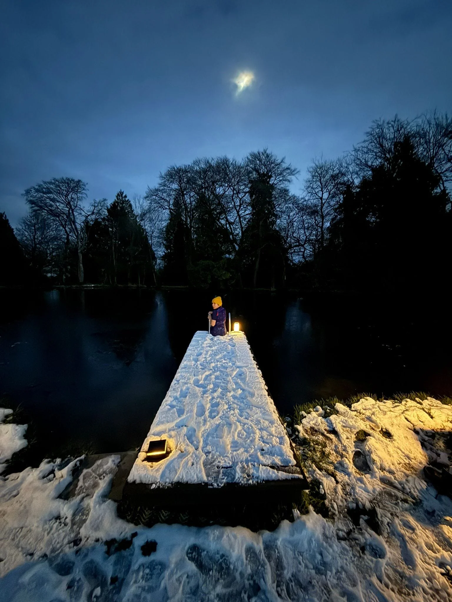 A person standing on a snow-covered pier by a dark river, with a full moon in the night sky and leafless trees in the background.