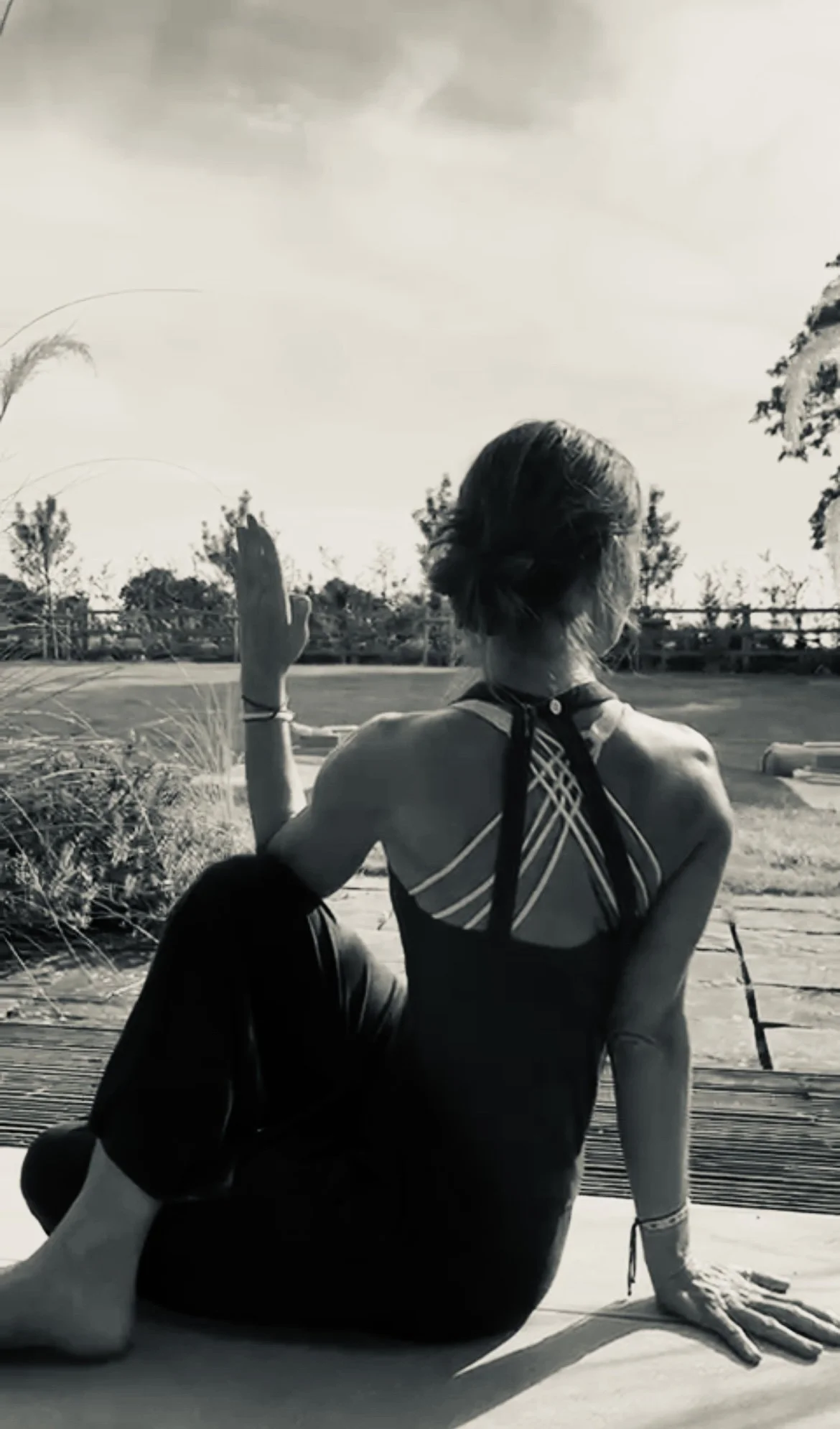 A woman sitting outdoors on a wooden deck, looking away from the camera, with her hair in a bun, wearing a strappy top, with trees and a fence in the background, under cloudy sky.