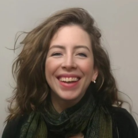 A smiling woman with brown, wavy hair, wearing earrings and a dark top, against a plain background.