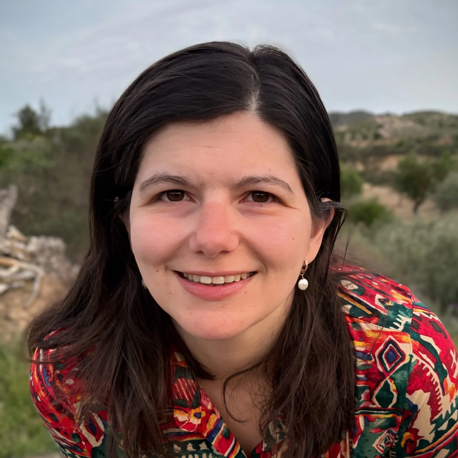 A woman with dark hair smiling outdoors with a natural landscape background.