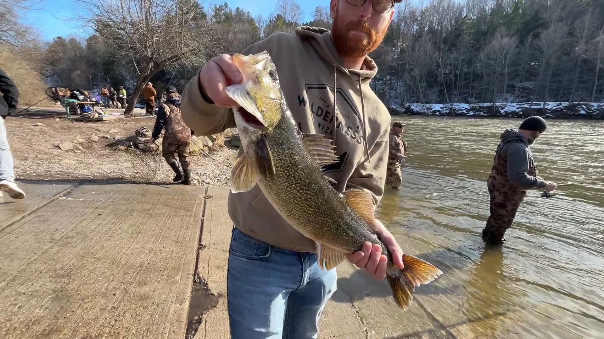Man holding a fish on a riverbank during a fishing trip with other anglers in the background.