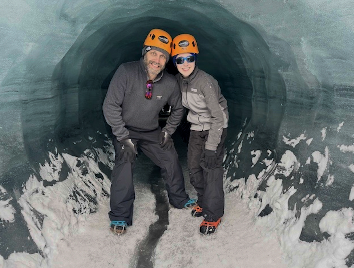 Two people, a man and a child, wearing helmets, gloves, and winter gear, are inside an ice tunnel with snow on the ground, smiling at the camera.