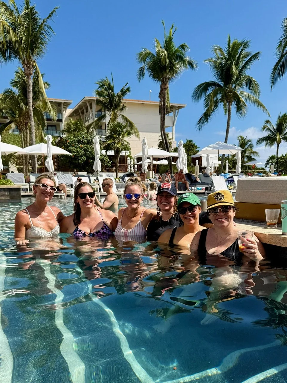Group of six women in swimsuits and sunglasses standing in a swimming pool at a tropical resort on a sunny day with palm trees and poolside lounge chairs visible in the background.