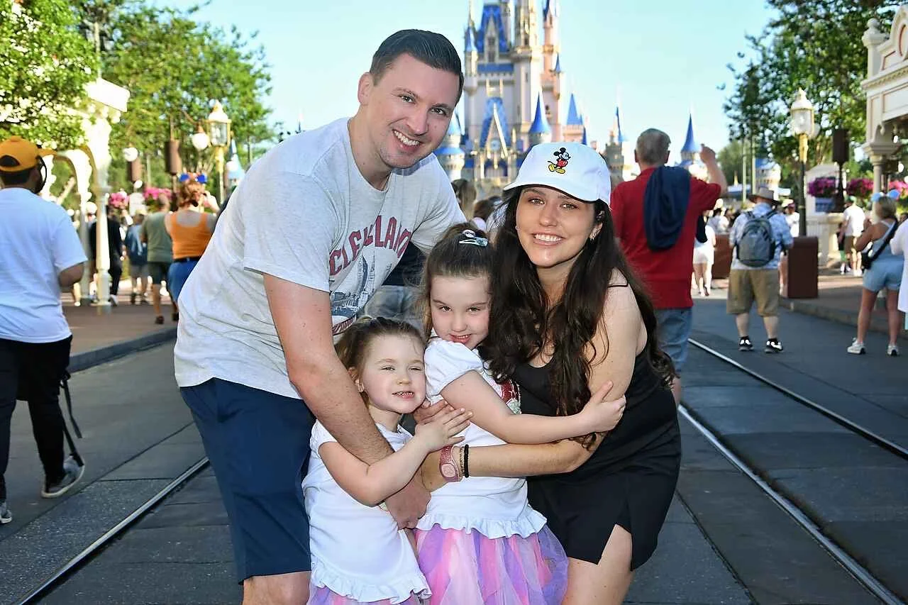 A happy family of four hugging in front of Sleeping Beauty Castle at Disneyland, with other park visitors in the background.