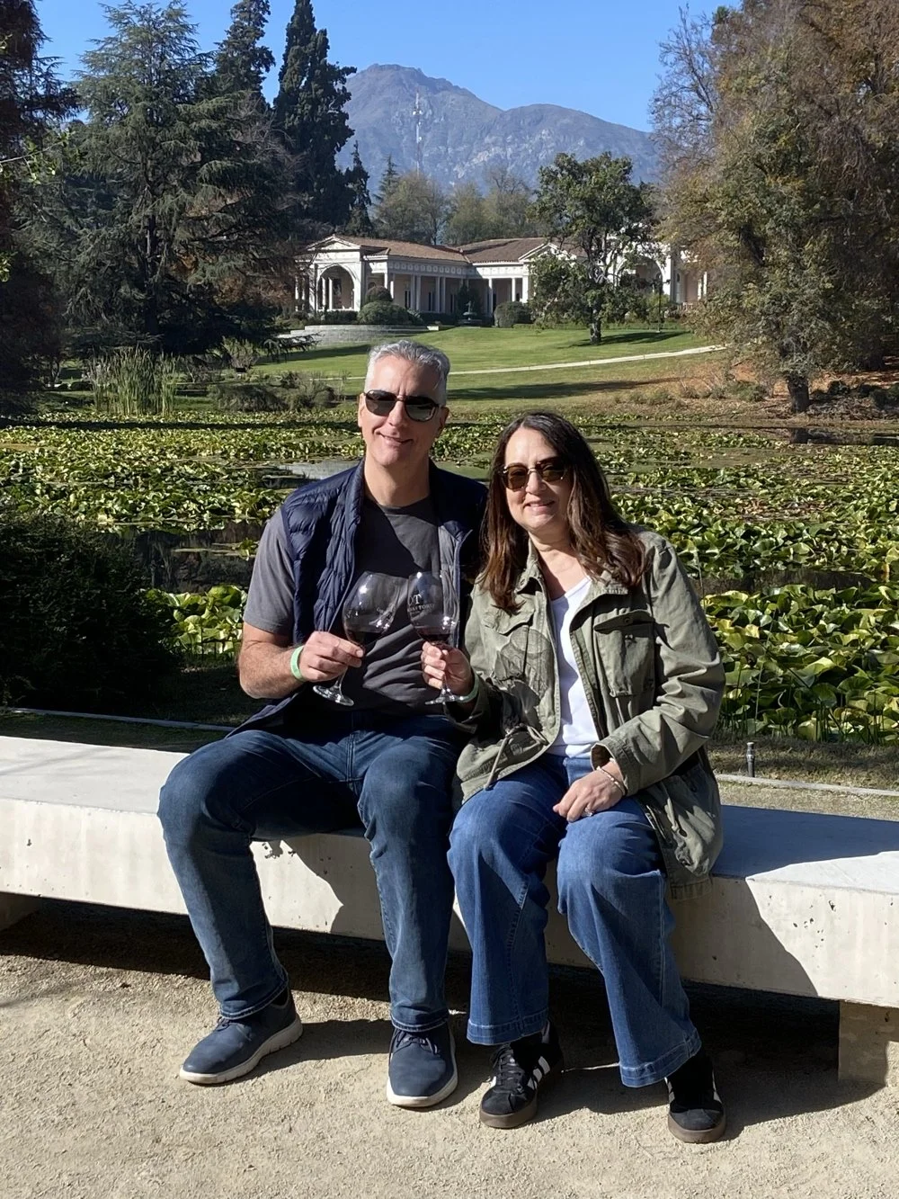 A couple holding wine glasses sitting on a concrete bench outdoors with pond and landscaped garden, trees, a grand house, and mountains in the background.