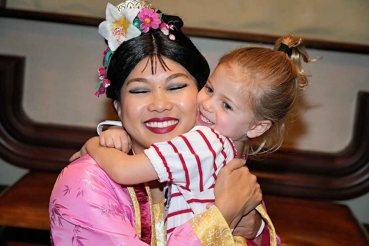 A woman dressed in traditional Asian attire and makeup is hugging a young girl with blond hair. The woman has an elaborate hairstyle decorated with flowers and wears bright pink clothing. The young girl is smiling and wearing a white shirt with red stripes.
