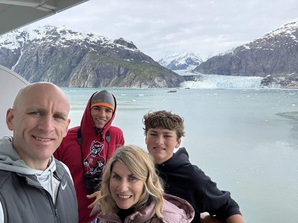 A family of four taking a selfie on a boat with a glacial fjord and snow-capped mountains in the background.