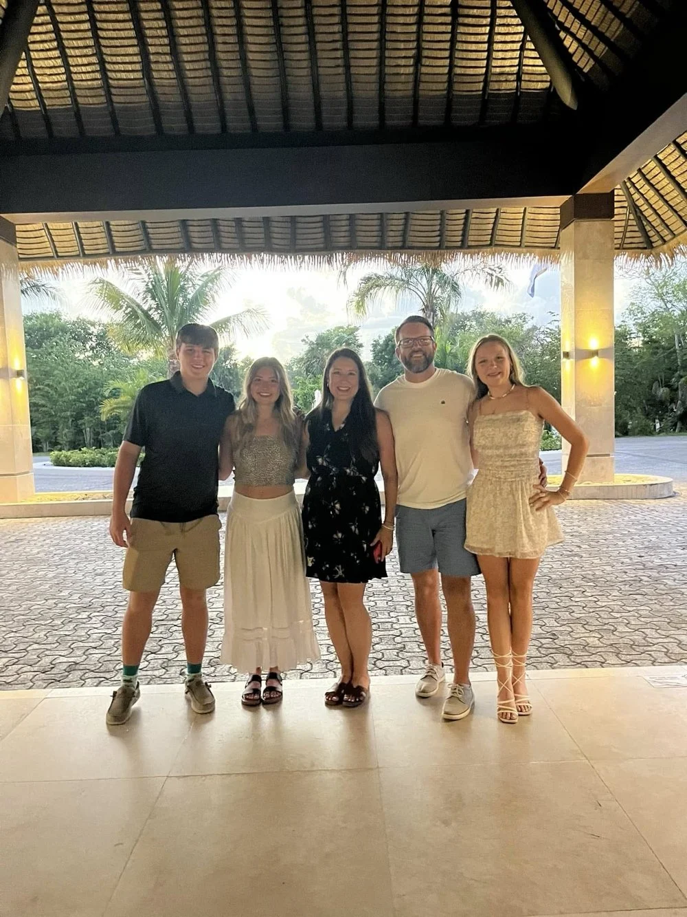 Group of five young adults standing together under a thatched roof, with palm trees and a sunset in the background.