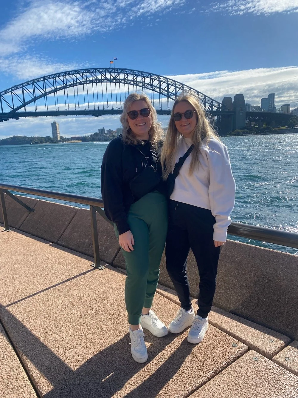 Two women standing on a waterfront promenade in front of the Sydney Harbour Bridge, smiling at the camera, with city buildings in the background.