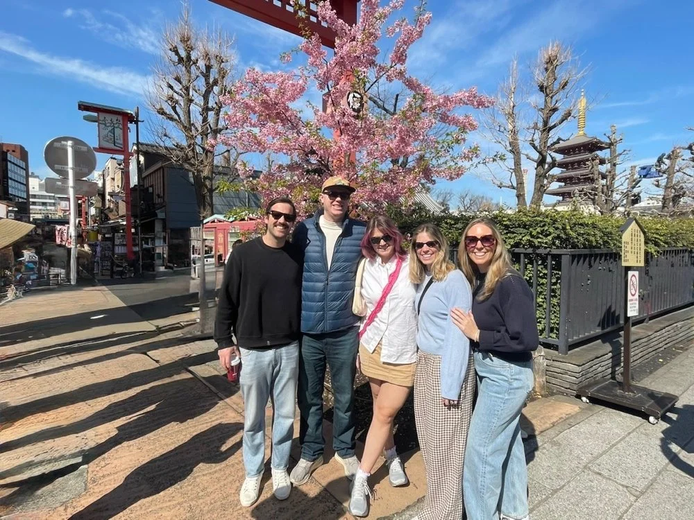 Group of five friends posing outdoors under a pink cherry blossom tree, with a traditional Japanese pagoda in the background on a sunny day.