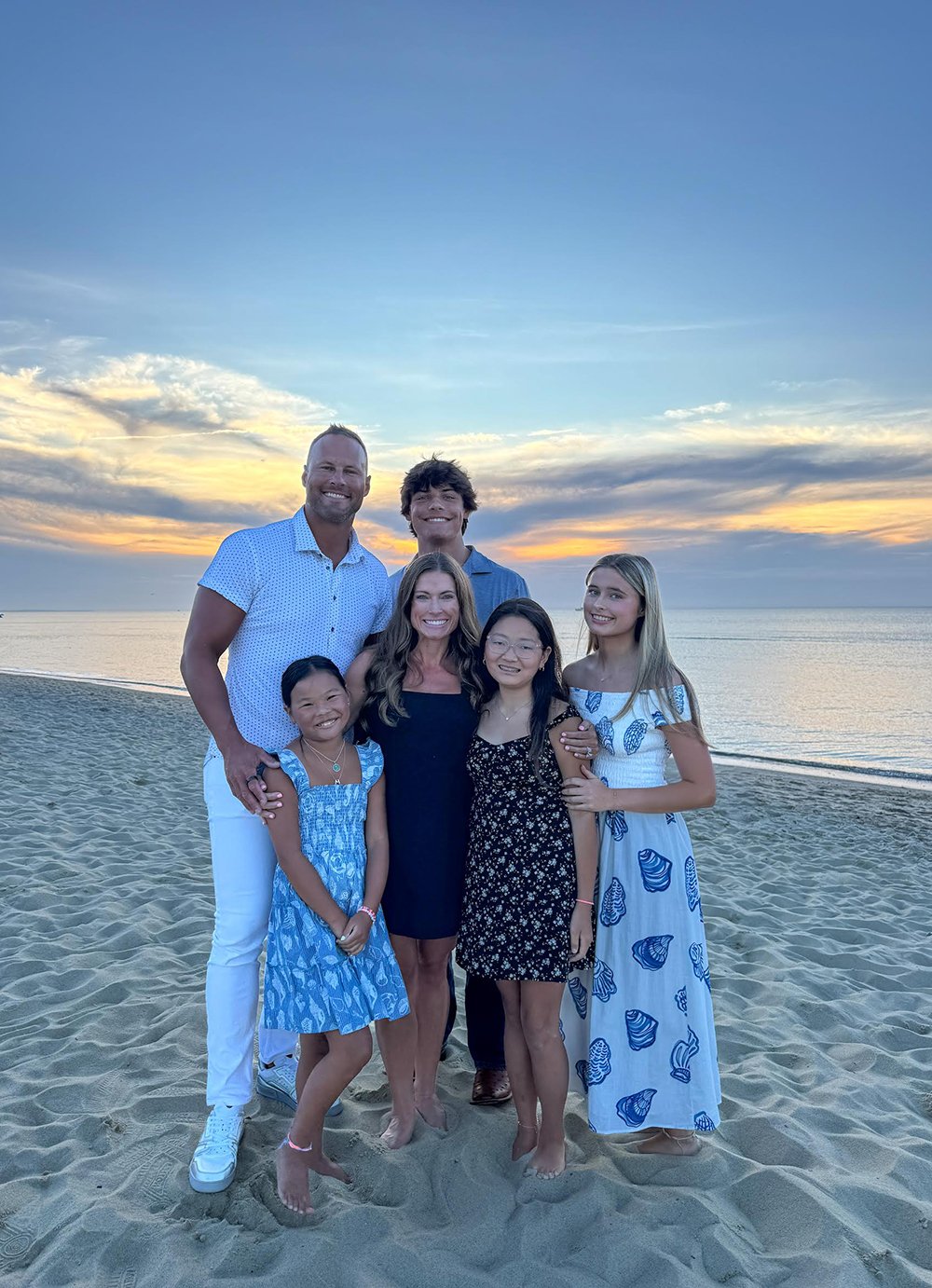 Shay's family standing on a sandy beach at sunset, smiling, with ocean and colorful sky in the background.