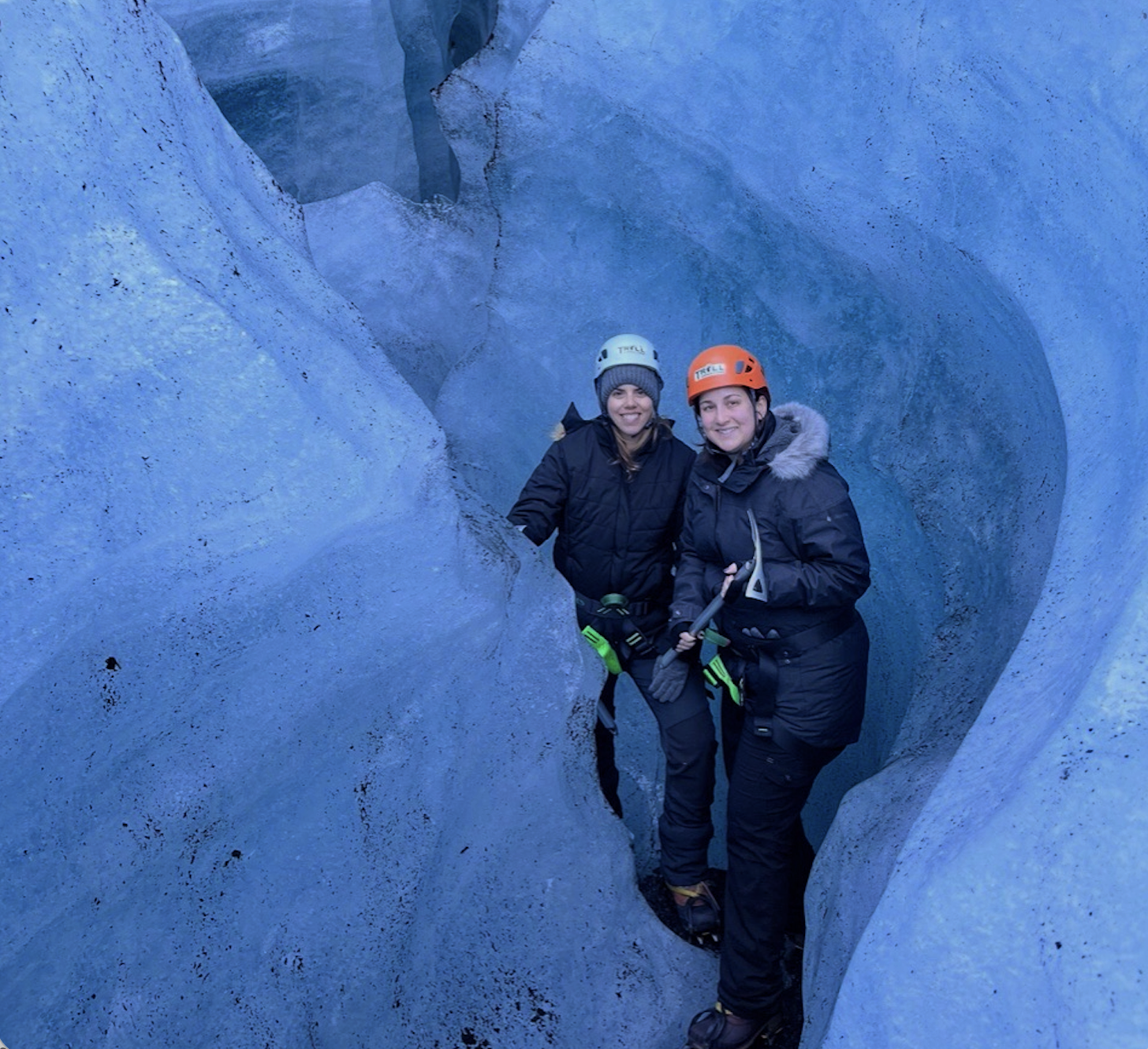 Two women wearing helmets and cold weather gear stand inside a narrow ice crevice, surrounded by blue ice walls, and smile at the camera.