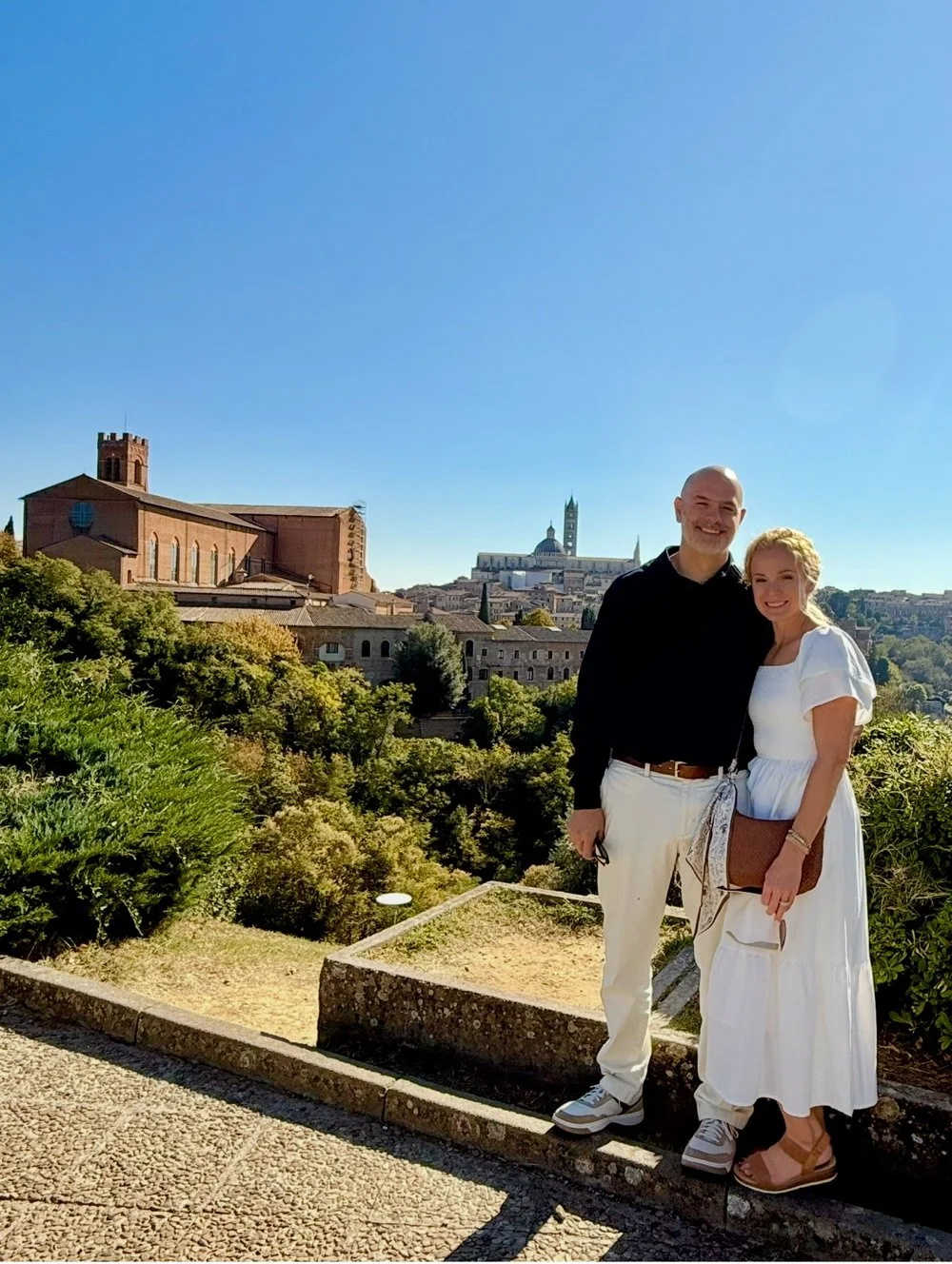 A smiling couple standing on a stone ledge with a historic European cityscape in the background, including a large church or cathedral with a tower, and lush greenery around them.
