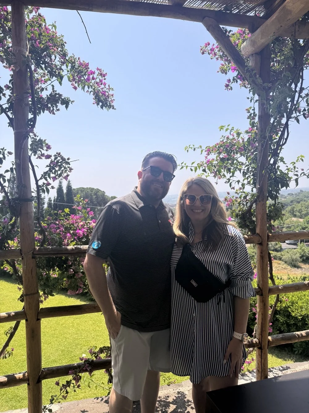 A man and woman smiling outdoors under a wooden arch with pink flowers, wearing sunglasses, in a sunny garden setting.