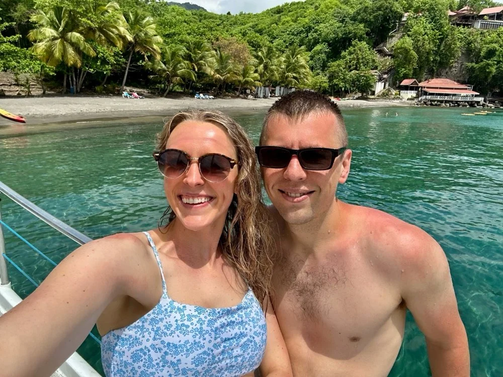 A smiling woman and man taking a selfie on a boat in a tropical location with clear water, beach, palm trees, and hillside buildings in the background.