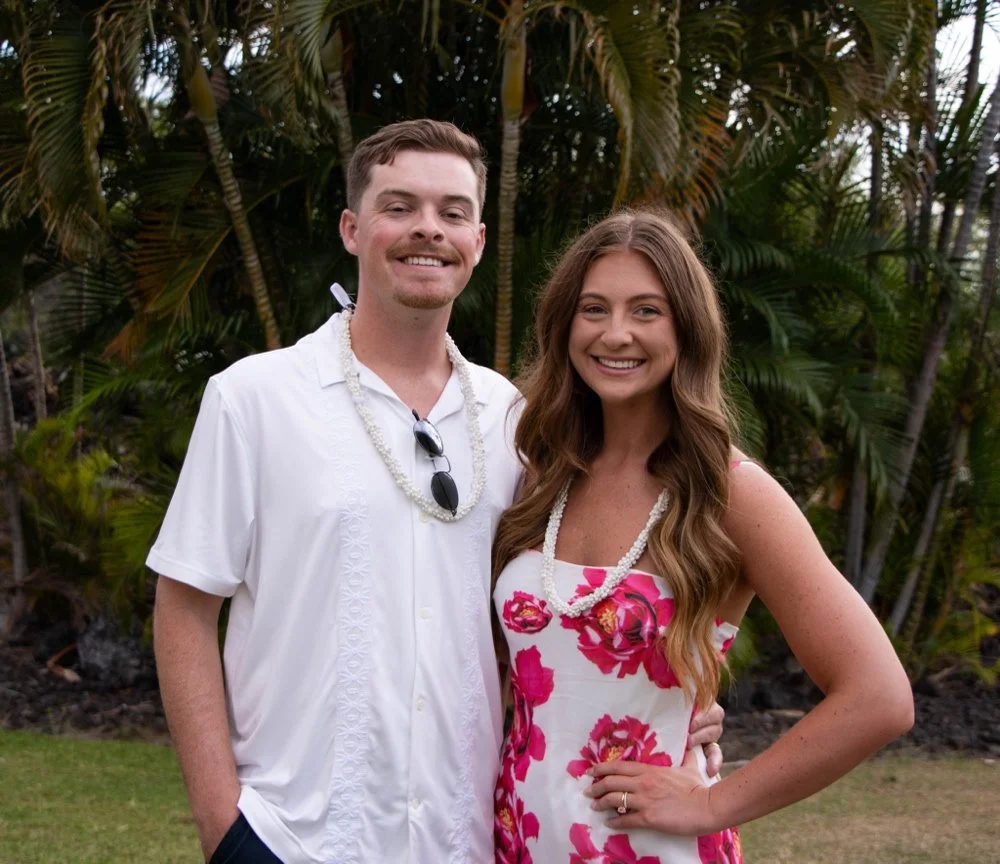 A smiling couple standing outdoors in front of tropical palm trees, with the man wearing a white button-up shirt and sunglasses hanging from his shirt and the woman wearing a floral dress with pearl necklaces.