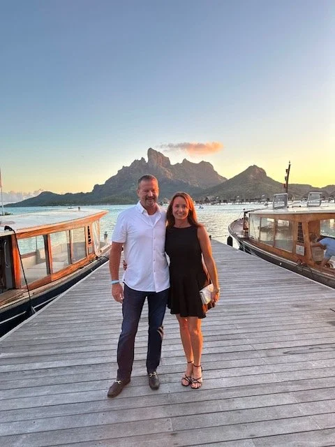 A man and woman standing on a wooden dock at sunset with boats and mountains in the background.