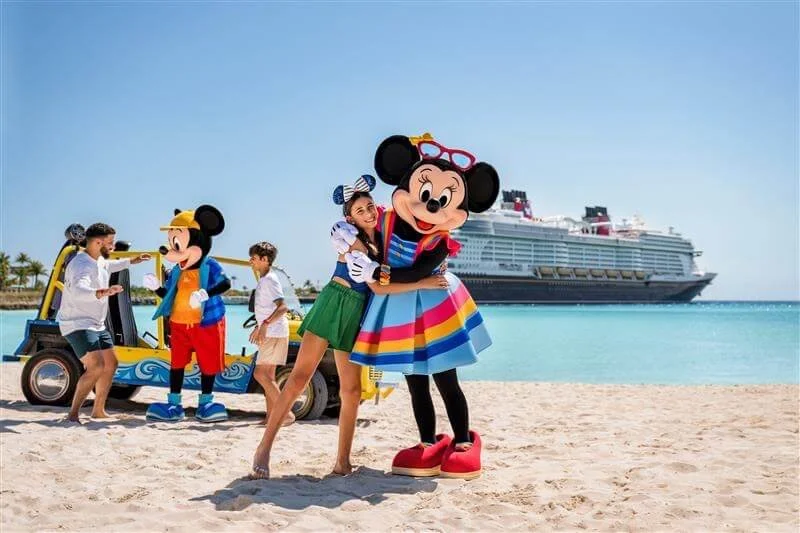 A woman hugging Mickey Mouse in a colorful dress on a beach with a cruise ship in the background, while three people and two additional Mickey Mouse characters are nearby.