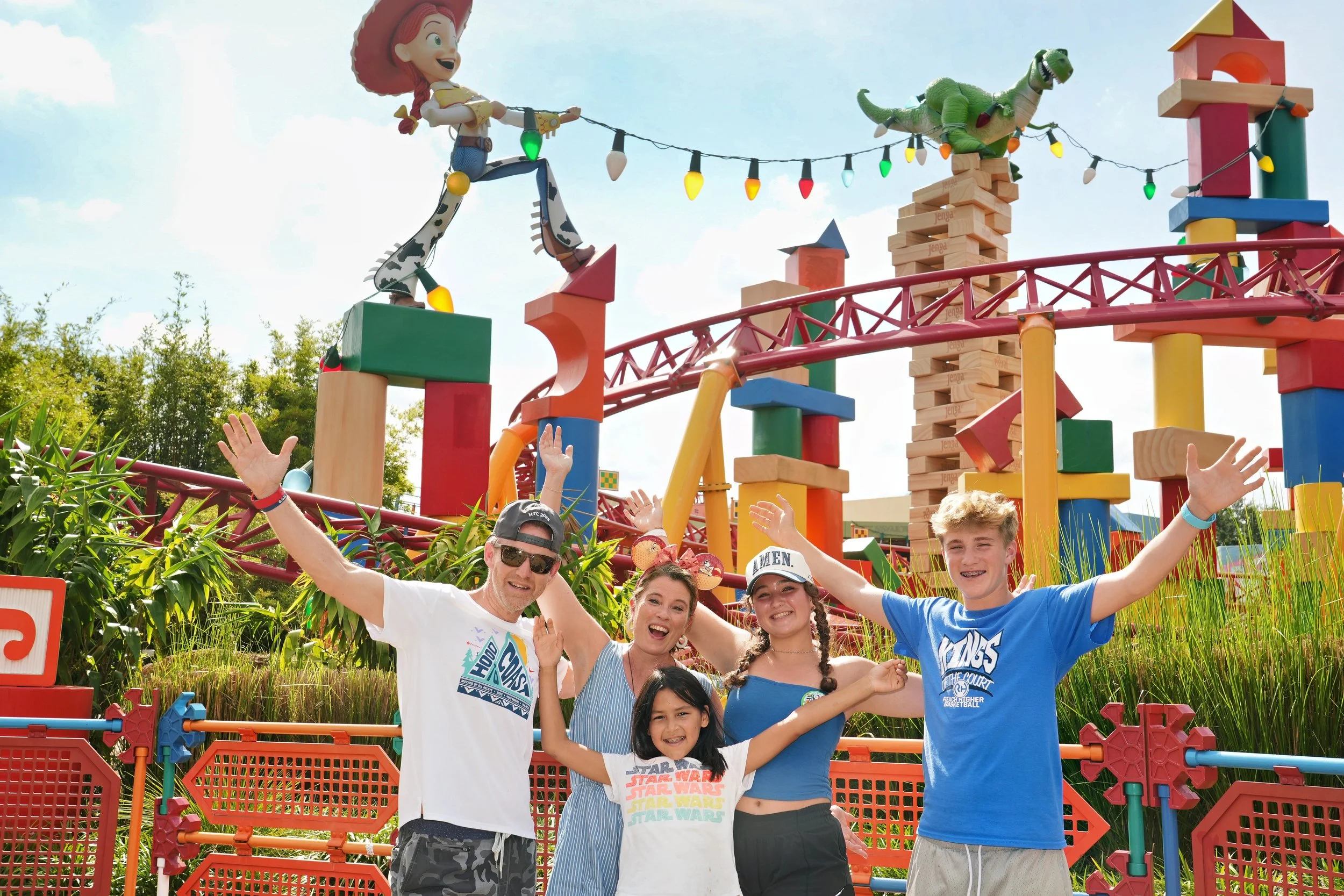 A group of five people, including three children and two adults, smiling and posing with their arms raised in front of a colorful amusement park ride featuring large characters of Sheriff Woody and Rex from Toy Story, with a blue sky and trees in the background.