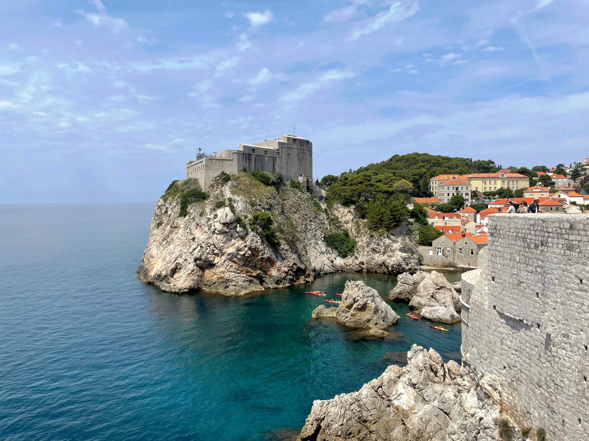 A historical stone fortress built on a rocky cliff overlooking the blue ocean, with colorful buildings and green trees nearby, under a partly cloudy sky, and people kayaking in the water.
