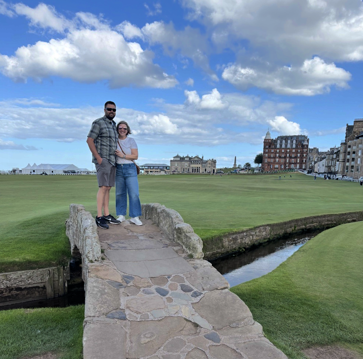 A man and woman standing on a small stone bridge on a golf course, with buildings and a cloudy sky in the background.