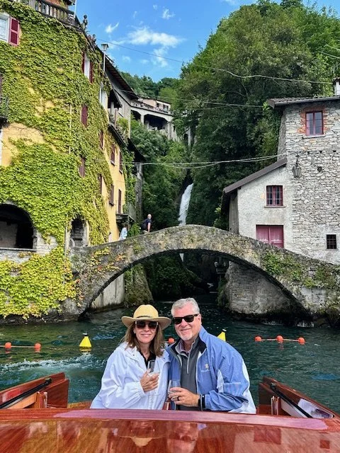 A couple sitting on a boat with the cityscape, stone bridge, waterfall, and greenery in the background.