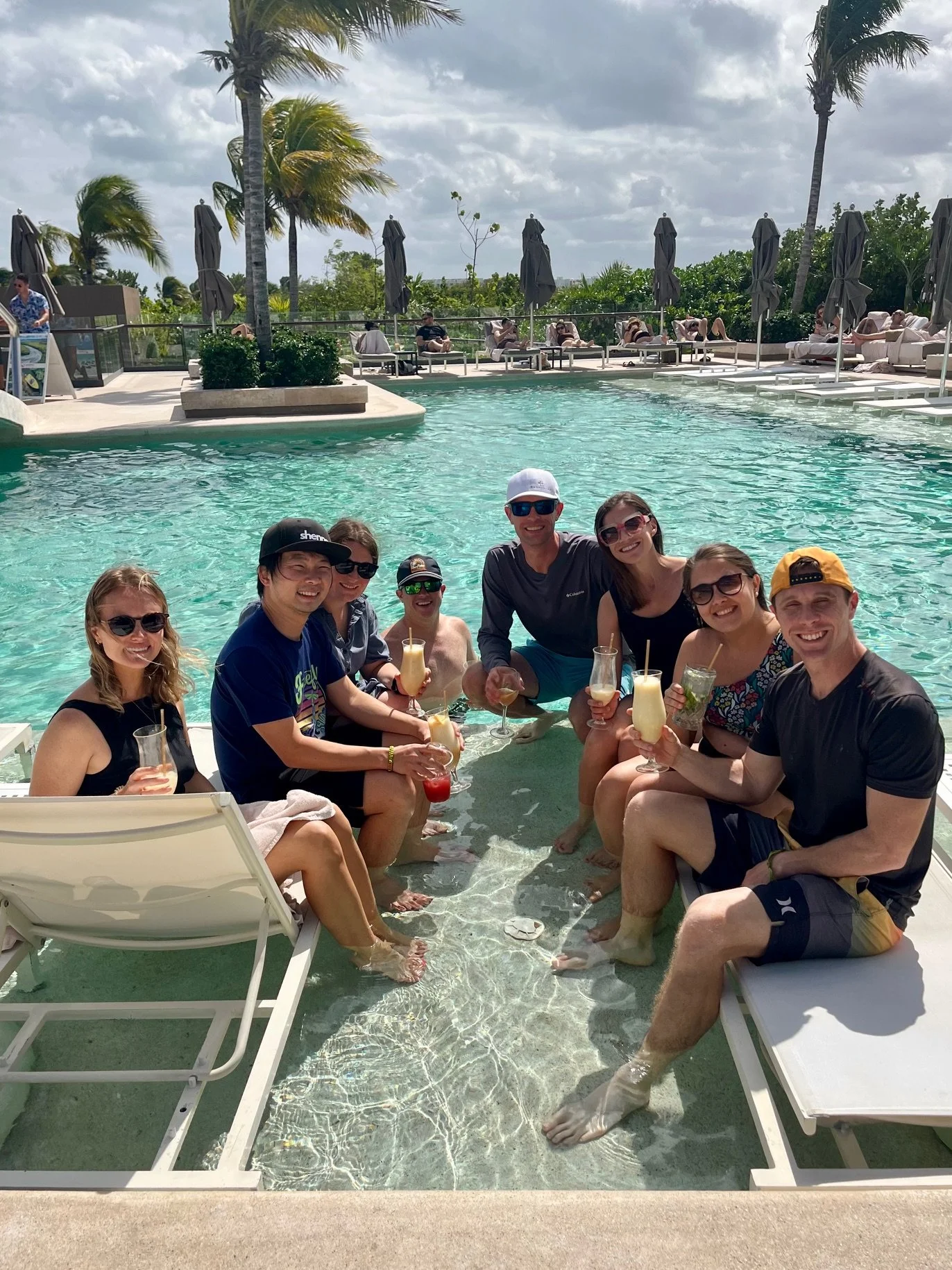 A group of nine people gathered in a pool, smiling and holding drinks. They are sitting partially in the water with some standing or sitting on the poolside. The background features lounge chairs, umbrellas, palm trees, and a partly cloudy sky.