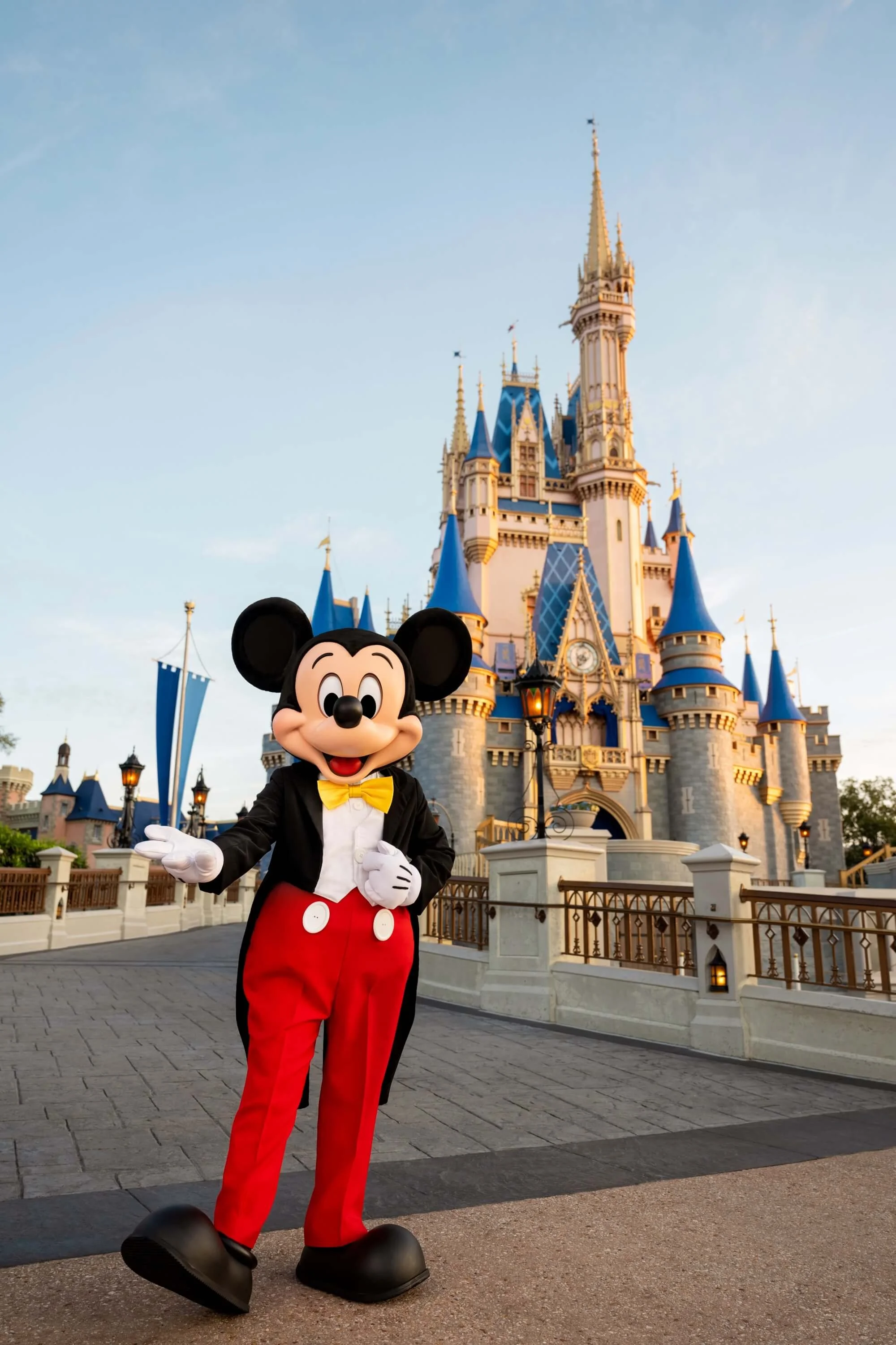 Mickey Mouse mascot standing in front of a Disney castle at Disney theme park during the day.