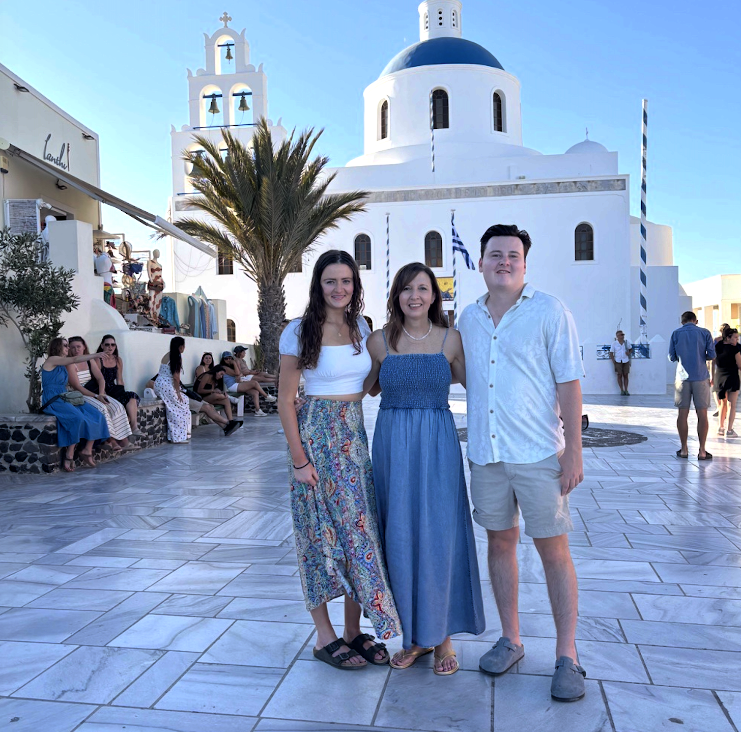 Three young adults stand together in front of a white chapel with a blue dome, with other people sitting on benches and walking nearby, in a sunny outdoor setting with a palm tree.